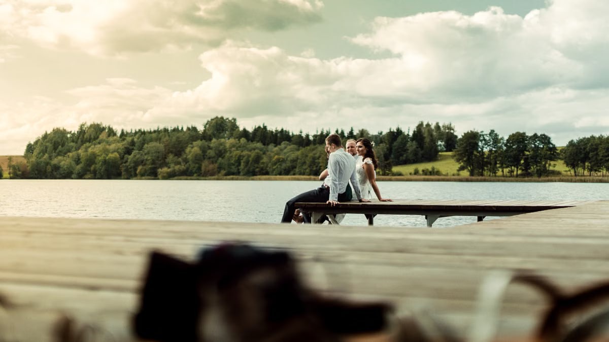 Three adults enjoy a serene moment sitting on a wooden dock overlooking a tranquil lake surrounded by greenery.